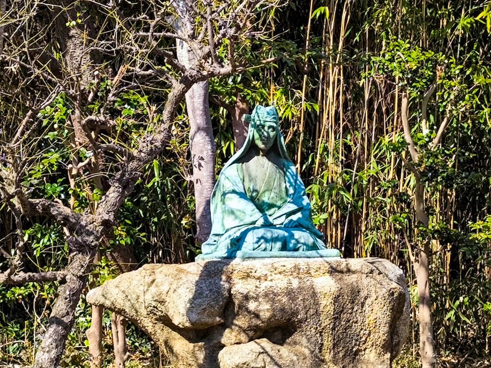 Die Bronzestatue einer sitzenden Figur in traditionellen Gewändern und mit Kopfbedeckung steht auf einem großen Stein in Okutsunomiya, umgeben von dichten Bäumen und Bambus in einer sonnenbeschienenen Außenanlage.