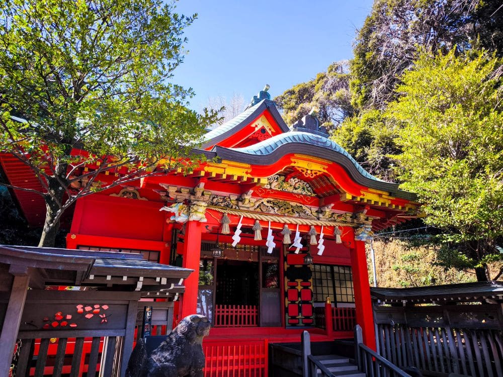 Ein leuchtend roter Nakatsunomiya Shinto-Schrein mit verschnörkelten Dächern steht umgeben von grünen Bäumen unter einem klaren blauen Himmel und schafft eine friedliche und traditionelle japanische Atmosphäre.