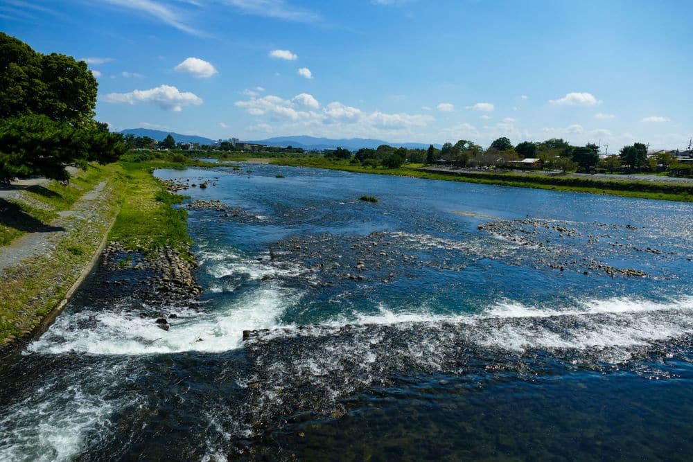 Ein breiter, sanft fließender Fluss mit kleinen weißen Stromschnellen schlängelt sich durch Arashiyama, gesäumt von grünem Gras und Bäumen unter einem strahlend blauen Himmel mit vereinzelten Wolken. Am Horizont sind Häuser und ferne Berge zu sehen.