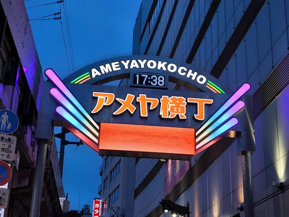 Das beleuchtete Eingangsschild von Ameya Yokocho leuchtet in bunten Lichtern unter dem Abendhimmel und zeigt japanische Schriftzeichen sowie eine Digitaluhr mit der Uhrzeit 17:38 Uhr. Im Hintergrund schlängelt sich die Yamanote-Linie durch die umliegenden Gebäude.