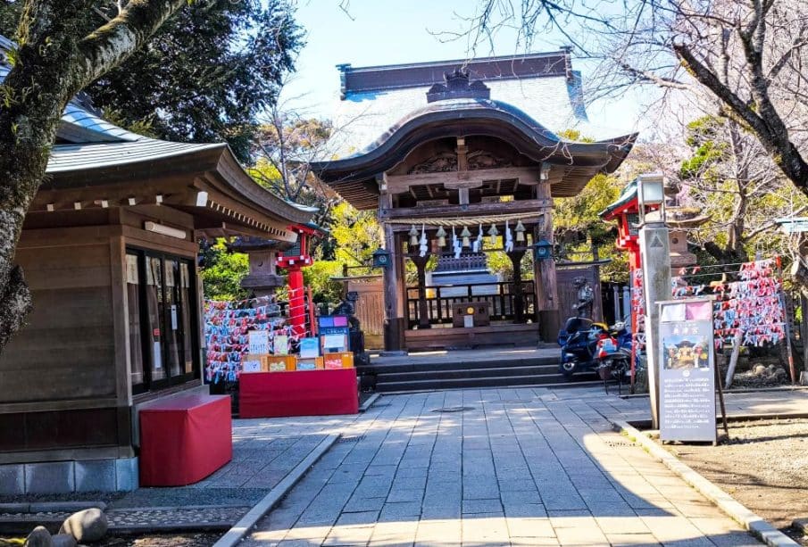 Ein Shinto-Schrein mit einem hölzernen Eingang, Laternen und an Ständern gebundenen Glückspapieren. Der Weg führt zum Hauptgebäude des Okutsunomiya-Schreins, das von Bäumen unter einem klaren blauen Himmel umgeben ist.