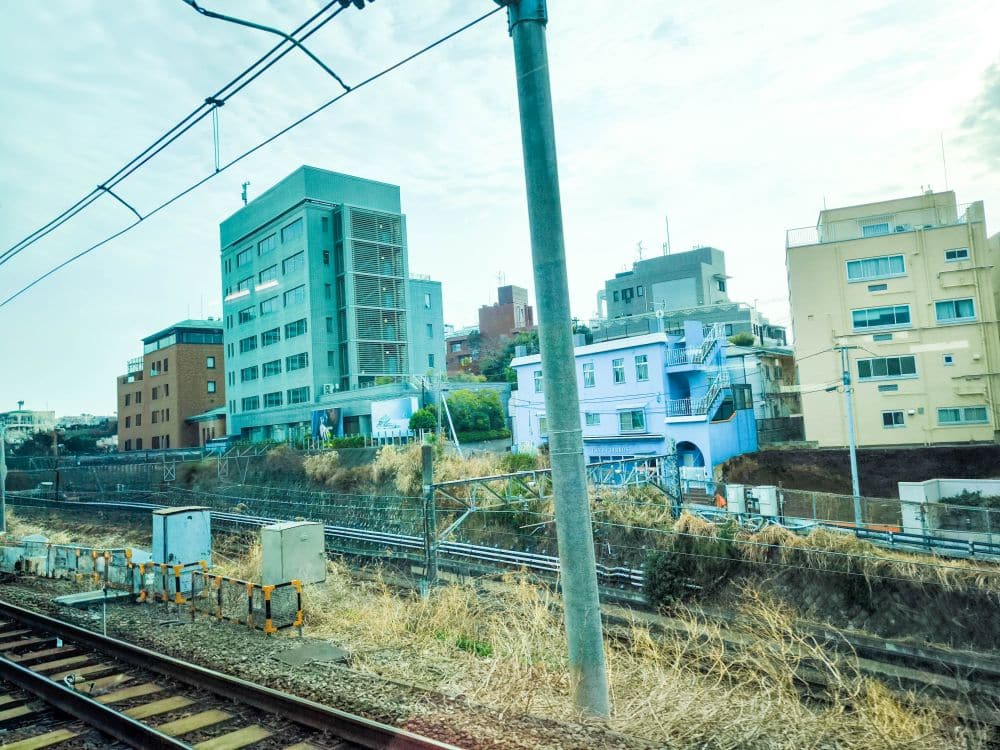 Aus dem Zugfenster betrachtet man die Yamanote-Linie mit ihren Gleisen, die von üppiger Grasfläche gesäumt sind. Im Hintergrund ragen mehrere Gebäude und Strommasten unter einem weiten blauen Himmel empor und bilden ein urbanes Bild.