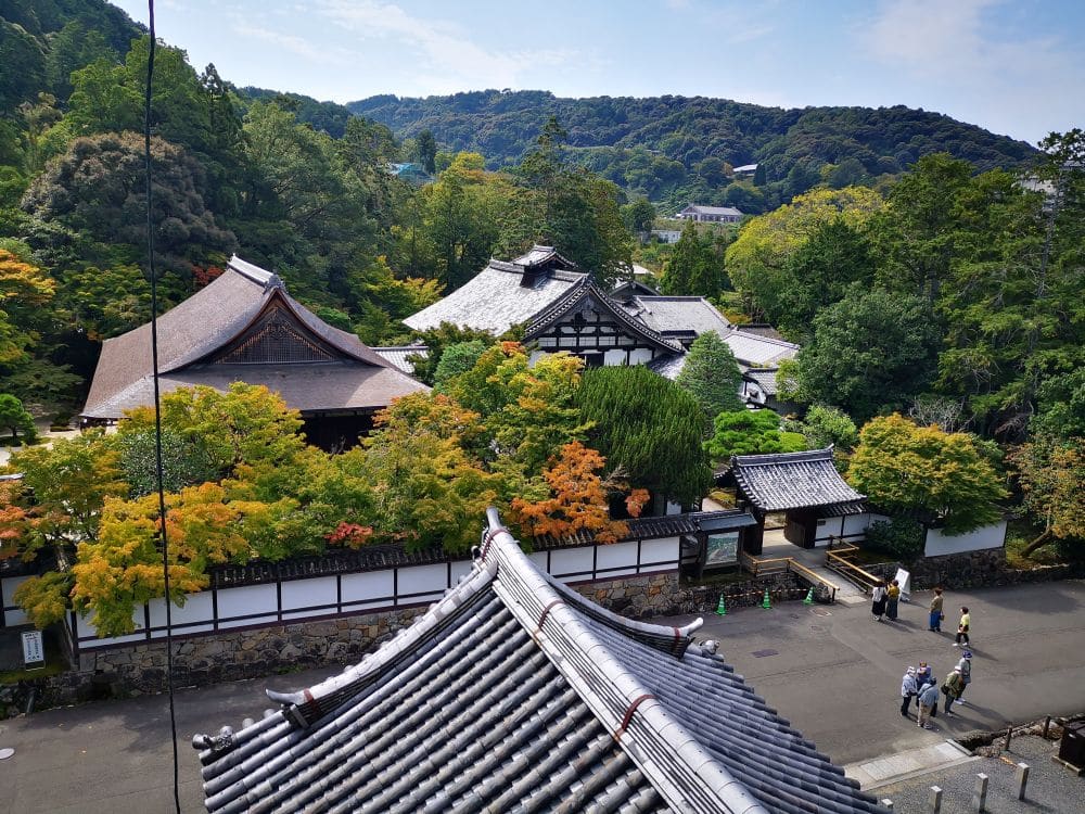 Luftaufnahme von traditionellen japanischen Tempelgebäuden mit einem mondähnlichen Eingangstor, umgeben von bunten Herbstbäumen und grünen Bergen, während ein paar Menschen in der Nähe spazieren gehen.