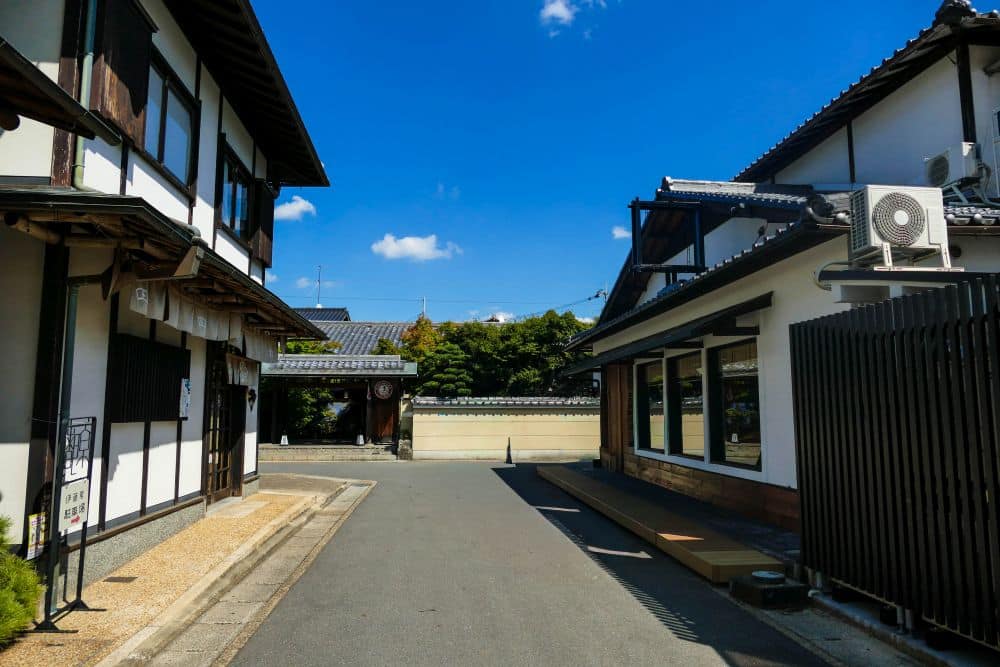 Eine ruhige Straße in Arashiyama mit traditionellen japanischen Gebäuden mit weißen Wänden und dunklen Holzakzenten unter einem klaren blauen Himmel. Im Hintergrund sind Bäume und eine Kachelwand zu sehen, die zu der ruhigen Atmosphäre beitragen.