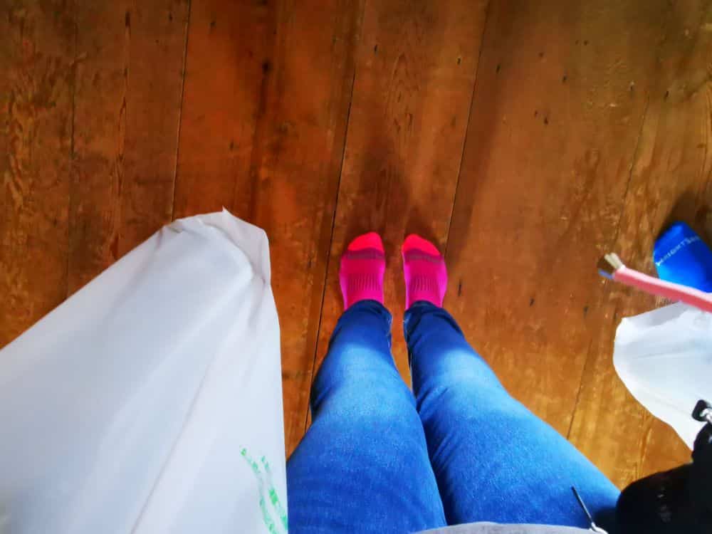 Ein Anfänger in hellrosa Socken und blauen Jeans steht auf einem Holzboden und hält eine weiße Plastiktüte in der Hand. Eine blaue Socke ist am rechten Bildrand teilweise sichtbar.