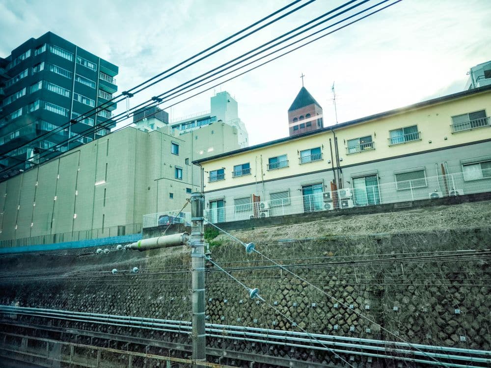 Blickt man aus dem Zugfenster, sieht man die Gleise der Yamanote-Linie. Inmitten der Szenerie ragt stolz ein hellbraunes Gebäude mit einem Kirchturm empor, der einen Kontrast zu einem modernen Wohnkomplex links bildet. Oberleitungen durchziehen elegant diese dynamische Stadtlandschaft.