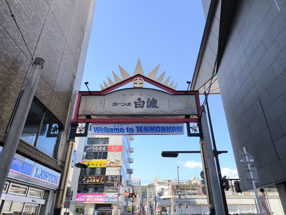 Ein Straßeneingang in Kagoshima mit einem Schild mit der Aufschrift Willkommen in TENMONKAN und japanischen Schriftzeichen, umgeben von hohen Gebäuden unter einem klaren blauen Himmel. Geschäfte und Straßenlaternen säumen den belebten Hintergrund.