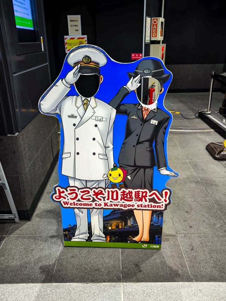 Eine farbenfrohe Kawagoe-Figur im Bahnhof Kawagoe zeigt einen männlichen und einen weiblichen Zugbegleiter mit Gesichtslöchern für Fotoaufnahmen. Der Text lautet: "Willkommen im Bahnhof Kawagoe!" in Japanisch und Englisch.