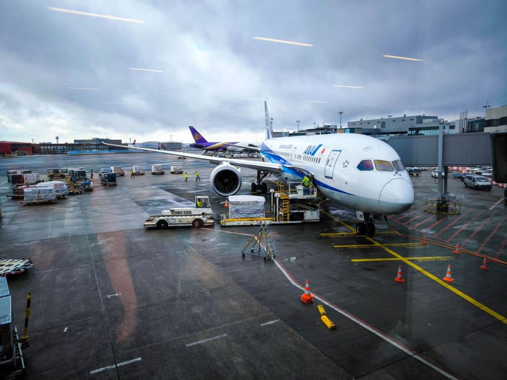 Ein großes Passagierflugzeug an einem Flugsteig wird mit Fracht beladen. Bodenfahrzeuge und Personal arbeiten in der Nähe - eine typische Szene für diejenigen, die Flüge nach Japan buchen möchten. Die nasse Landebahn spiegelt den bedeckten Himmel wider.