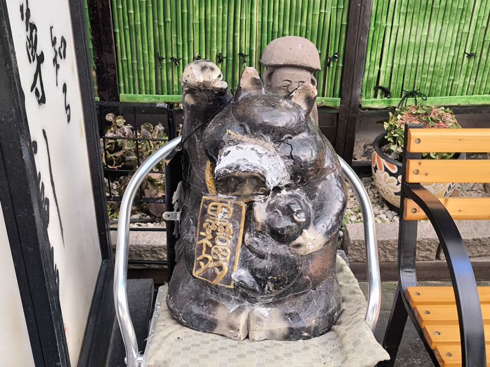 Eine verwitterte Marderhund-Statue, die ein goldenes Schild hält, sitzt auf einem Metallstuhl im Freien, neben einer gelb-schwarzen Bank und einem Bambuszaun - ein auffälliger Ort, der oft bei den geführten Touren in Kagoshima zu sehen ist. Der runde Bauch der Statue ist teilweise abgeplatzt.