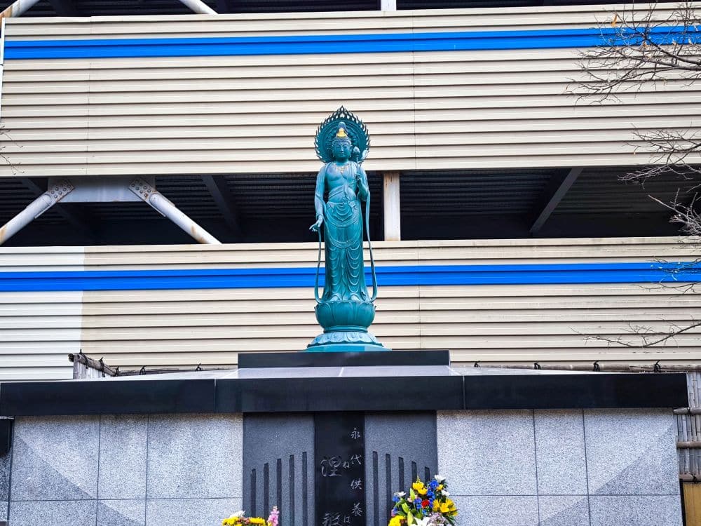 Eine grünlich-blaue Statue eines stehenden Buddhas steht auf einem Steinsockel in Kawagoe, auf dem bunte Blumen geopfert werden. Im Hintergrund ist ein modernes Gebäude mit horizontalen beigen und blauen Streifen zu sehen.