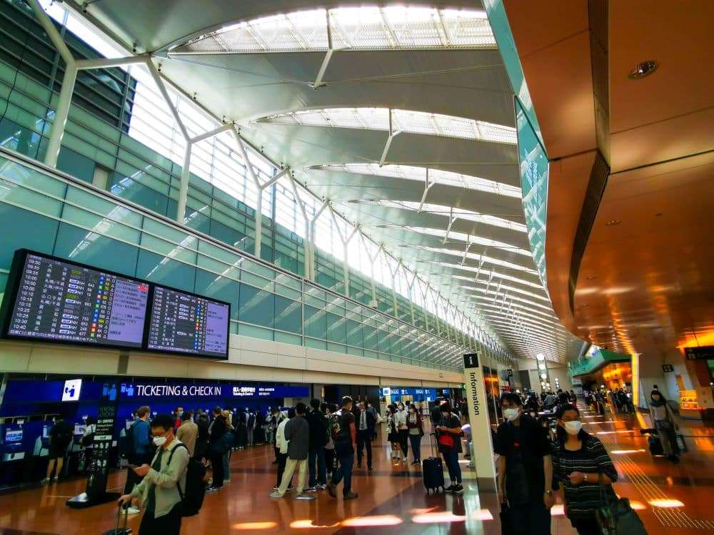Ein geschäftiges Haneda-Flughafenterminal mit Reisenden, von denen einige Masken tragen, die in der Nähe von Ticket- und Check-in-Schaltern gehen und warten. Große digitale Fluginformationstafeln sind über den Schaltern unter einer modernen, hellen Decke zu sehen.