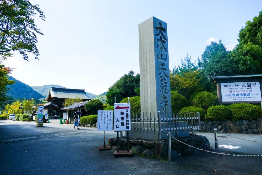 Ein hohes Steinmonument mit japanischen Inschriften steht am Eingang des Tenryu-ji-Tempels in Arashiyama, Kyoto, umgeben von Bäumen, Schildern und einem traditionellen Gebäude im Hintergrund unter einem blauen Himmel.