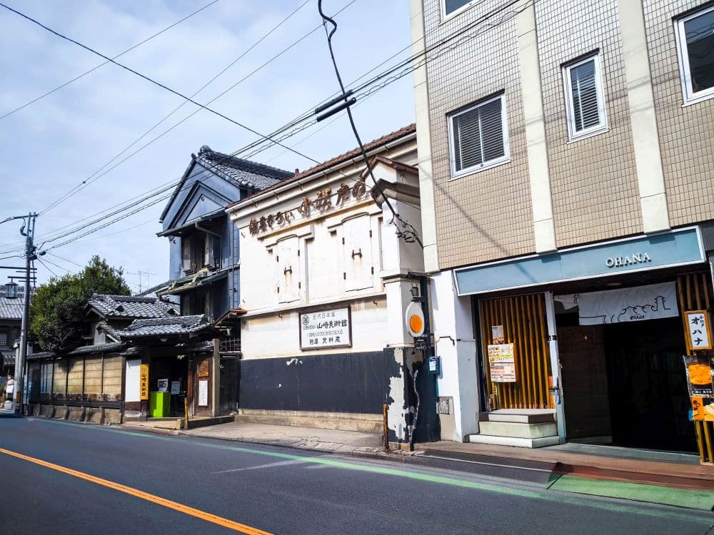 In einer ruhigen Straße in Kawagoe, Japan, stehen traditionelle und moderne Gebäude - ein weiß gemauertes Lagerhaus und ein Haus mit Ziegeldach neben einem beigen Gebäude mit der Aufschrift OHANA - während Oberleitungen den blauen Himmel durchkreuzen.