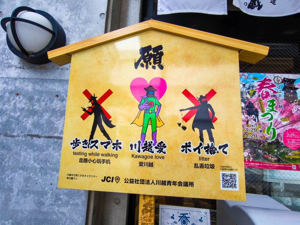 Auf einem gelben Schild in Kawagoe, das die Form einer Schreintafel hat, sind Cartoon-Figuren abgebildet, die davor warnen, beim Gehen SMS zu schreiben, in der Öffentlichkeit Zuneigung zu zeigen und Abfall zu entsorgen - jeweils mit einem roten X. Der Text ist auf Englisch und Japanisch abgedruckt.