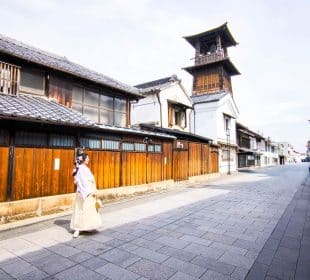 Eine Person in traditioneller japanischer Kleidung geht eine ruhige, gepflasterte Straße in Kawagoe entlang, die von Holzhäusern und dem hohen Uhrenturm Toki no Kane gesäumt ist, und das bei strahlendem Himmel.