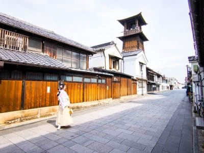 Eine Person in traditioneller japanischer Kleidung geht eine ruhige, gepflasterte Straße in Kawagoe entlang, die von Holzhäusern und dem hohen Uhrenturm Toki no Kane gesäumt ist, und das bei strahlendem Himmel.