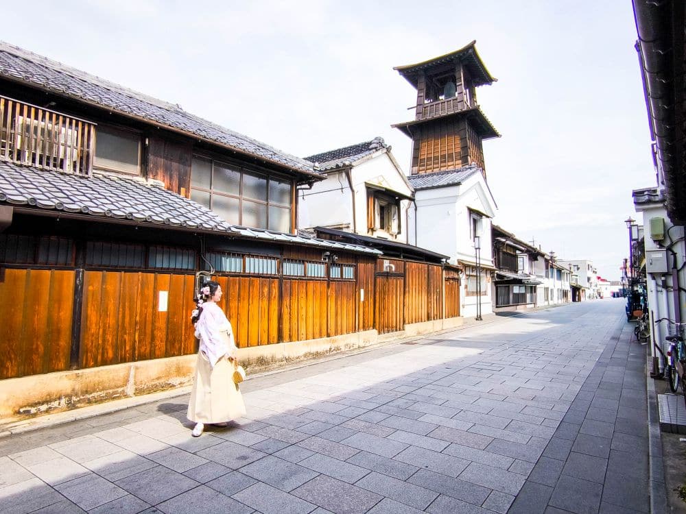 Eine Person in traditioneller japanischer Kleidung geht eine ruhige, gepflasterte Straße in Kawagoe entlang, die von Holzhäusern und dem hohen Uhrenturm Toki no Kane gesäumt ist, und das bei strahlendem Himmel.