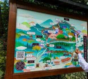 Eine große illustrierte Karte mit farbenfrohen Zeichnungen und japanischem Text zeigt eine Wanderroute durch bewaldete Berge, Schreine und Sehenswürdigkeiten; ein Tourguide zeigt auf die Karte, während eine andere Person zuschaut.