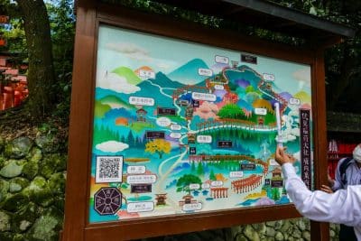 Eine große illustrierte Karte mit farbenfrohen Zeichnungen und japanischem Text zeigt eine Wanderroute durch bewaldete Berge, Schreine und Sehenswürdigkeiten; ein Tourguide zeigt auf die Karte, während eine andere Person zuschaut.