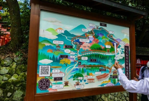 Eine große illustrierte Karte mit farbenfrohen Zeichnungen und japanischem Text zeigt eine Wanderroute durch bewaldete Berge, Schreine und Sehenswürdigkeiten; ein Tourguide zeigt auf die Karte, während eine andere Person zuschaut.