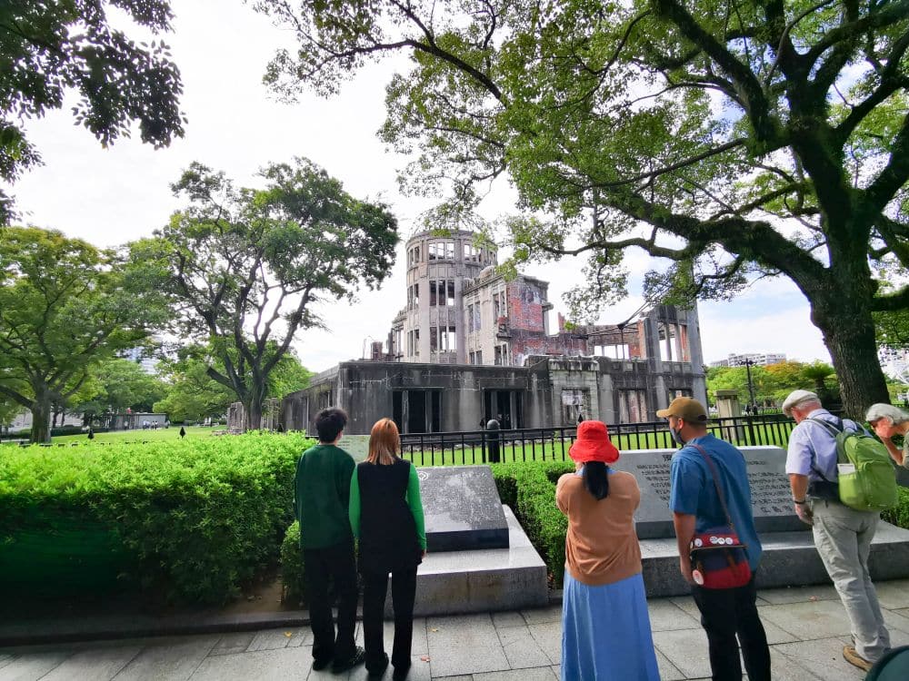 An einem sonnigen Tag stehen die Menschen vor dem Hiroshima-Friedensdenkmal (Atombombenkuppel), umgeben von grünen Bäumen und Sträuchern, während ein Tourguide Einblicke gewährt, während die Besucher Tafeln lesen und über die historische Stätte nachdenken.