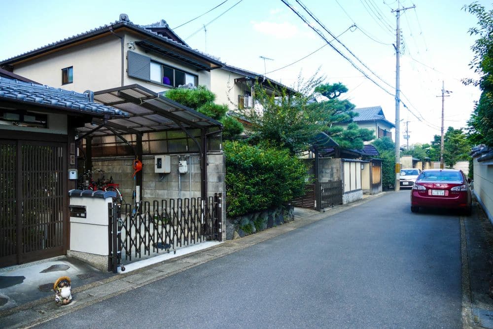 Eine ruhige Wohnstraße in Arashiyama, Japan, mit Häusern im traditionellen Stil, einem roten Auto auf der rechten Seite, Grün entlang der Zäune, Oberleitungen und einer Katze, die in der Nähe eines Tores im Vordergrund sitzt.