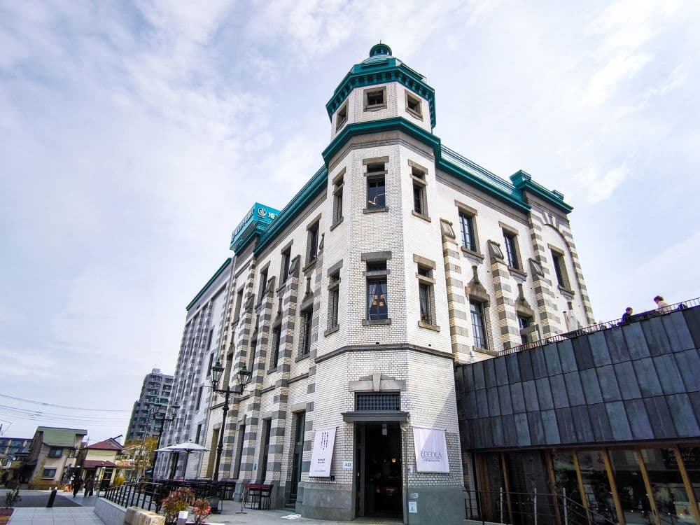 Ein großes, historisches weißes Backsteingebäude mit grünen Verzierungen und einem markanten Eckturm steht unter einem teilweise bewölkten Himmel in Kawagoe. In der Nähe gehen Menschen spazieren, und im Hintergrund sind moderne Gebäude zu sehen.
