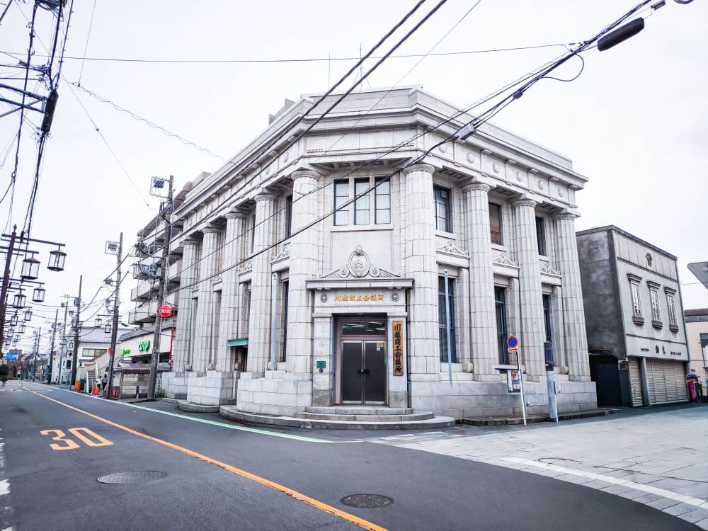 Ein großes, altmodisches Steingebäude mit Säulen und verzierten Details steht an einer Straßenecke in Kawagoe, Japan; darüber kreuzen sich Stromleitungen, und in einer ruhigen, leeren Straße sind nahe gelegene Geschäfte zu sehen.