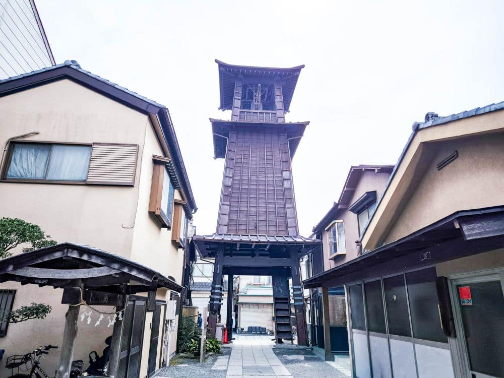 Der hohe, traditionelle japanische Uhrenturm aus Holz, Toki no Kane, steht zwischen zwei beigen Häusern mit Ziegeldächern an einem schmalen Steinweg in Kawagoe unter einem hellen, bewölkten Himmel.