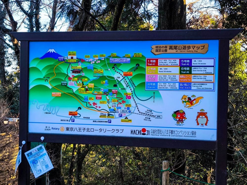 Eine farbenfrohe Wanderkarte von Takao in Japan ist im Freien ausgestellt und zeigt verschiedene Wanderrouten, Sehenswürdigkeiten und Einrichtungen, mit Cartoon-Maskottchen und dem Berg Fuji im Hintergrund. Es sind japanische und einige englische Texte zu sehen.