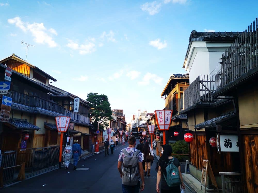 Die Menschen schlendern eine traditionelle japanische Straße entlang, die von Holzhäusern und Papierlaternen gesäumt ist, und werden von einem Tourguide unter einem blauen Himmel mit vereinzelten Wolken geführt. Die Atmosphäre ist ruhig und malerisch.