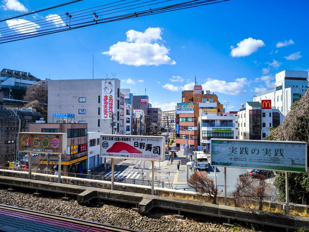 Blick vom Bahnsteig des Bahnhofs Hino auf eine japanische Stadtstraße mit bunten Gebäuden, Reklametafeln und Schildern - darunter ein McDonalds - unter einem strahlend blauen Himmel, der von vereinzelten Wolken durchzogen ist.