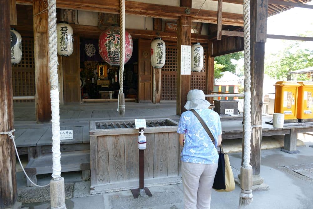 Eine ältere Person mit Hut und blauem Hemd steht vor einem hölzernen Schrein und scheint zu beten, während ein Fremdenführer Einblicke gewährt. Große weiße Seile, Papierlaternen und Holzkonstruktionen sind zu sehen, und das Sonnenlicht beleuchtet die Szene.