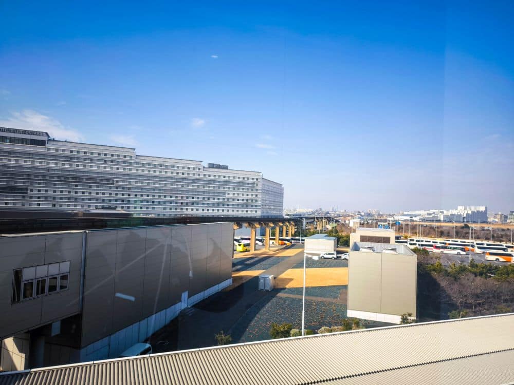 Ein heller, klarer Tag in Haneda mit einem großen modernen Gebäude auf der linken Seite, einer gepflasterten Straße mit geparkten Bussen in der Mitte und weiteren Gebäuden und Grünflächen im Hintergrund unter einem strahlend blauen Himmel.