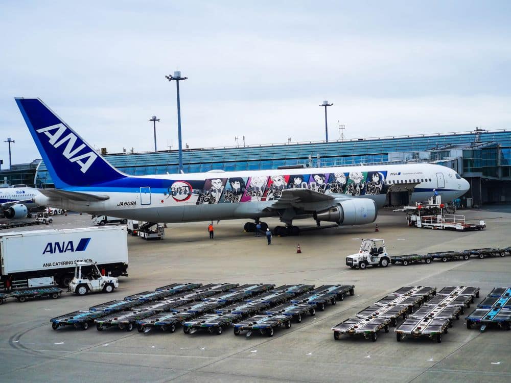 Ein großes ANA-Flugzeug mit bunten Zeichentrickfiguren auf der Seite ist an einem Flughafengate geparkt und lädt Reisende ein, Flüge nach Japan in Betracht zu ziehen, während in der Nähe Fahrzeuge des Bodenservice und leere Gepäckwagen warten.