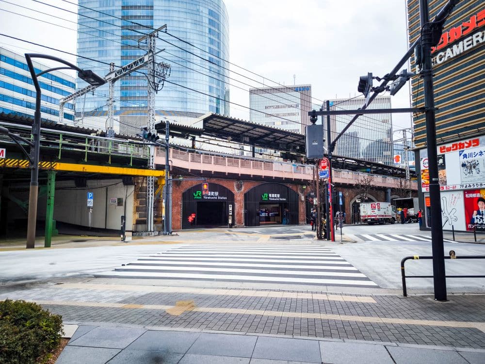 Straßenansicht eines japanischen Stadtgebiets mit den Hochgleisen der Yamanote-Linie, einem Fußgängerüberweg, Wolkenkratzern und Ladenschildern. Der klare Himmel und die japanischen Schilder tragen zur geschäftigen Stadtatmosphäre bei.