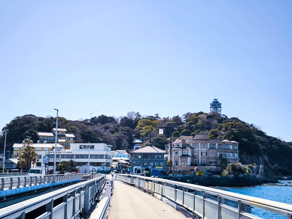 Ein gepflasterter Weg mit Geländer führt über das Wasser zu einer Ansammlung von Gebäuden und üppigen, grünen Hügeln. Der ikonische Aussichtsturm Enoshima Sea Candle erhebt sich über den Bäumen unter einem klaren blauen Himmel.