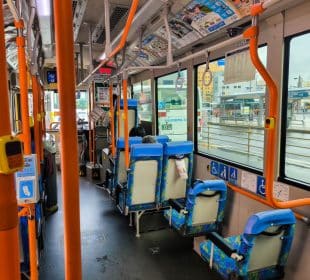 Innenraum eines Stadtbusses mit blauen und orangefarbenen Sitzen, Handläufen und Hinweisschildern für Barrierefreiheit. Nur wenige Fahrgäste sitzen. Große Fenster zeigen einen regnerischen Tag draußen und eine nahegelegene Bushaltestelle oder einen Bahnhof im Hintergrund.