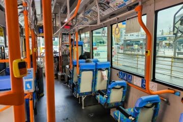 Innenraum eines Stadtbusses mit blauen und orangefarbenen Sitzen, Handläufen und Hinweisschildern für Barrierefreiheit. Nur wenige Fahrgäste sitzen. Große Fenster zeigen einen regnerischen Tag draußen und eine nahegelegene Bushaltestelle oder einen Bahnhof im Hintergrund.
