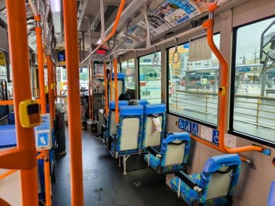Innenraum eines Stadtbusses mit blauen und orangefarbenen Sitzen, Handläufen und Hinweisschildern für Barrierefreiheit. Nur wenige Fahrgäste sitzen. Große Fenster zeigen einen regnerischen Tag draußen und eine nahegelegene Bushaltestelle oder einen Bahnhof im Hintergrund.