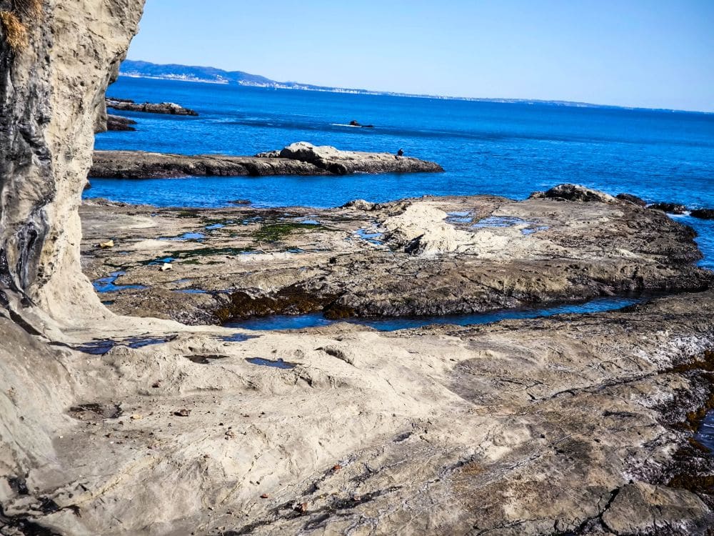 Felsige Iwaya-Küste mit unebener, strukturierter Oberfläche neben klarem, blauem Meerwasser unter hellem Himmel. Am Horizont sind weit entfernte Landformen über dem Wasser zu sehen. Im unmittelbaren Vordergrund sind keine Menschen zu sehen.