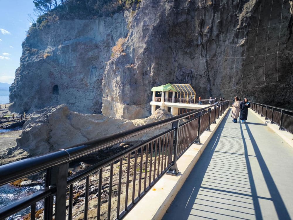 Die Menschen gehen auf einem gepflasterten Weg mit schwarzem Geländer neben der Iwaya-Felsklippe entlang, mit einer kleinen Schutzhütte vor sich und dem Meer auf der linken Seite unter einem klaren blauen Himmel.