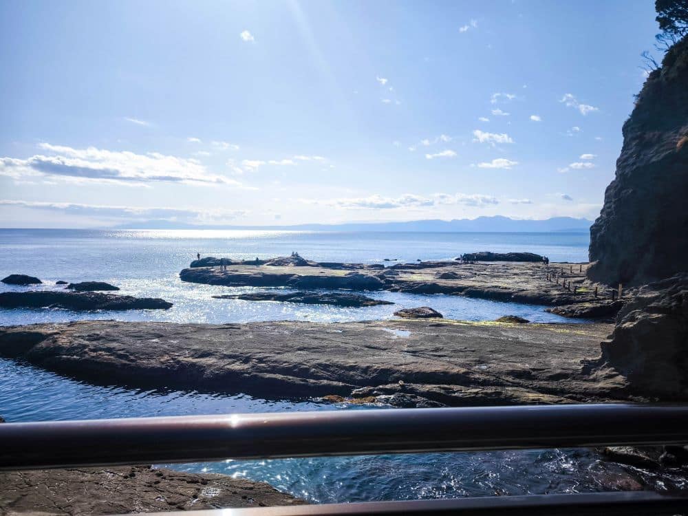 Felsenküste unter strahlend blauem Himmel, wobei sich das Sonnenlicht auf dem ruhigen Meer bei Iwaya spiegelt. In der Ferne stehen Silhouetten von Menschen auf den Felsen, am Horizont tauchen Landformen auf, und im Vordergrund ist ein Geländer zu sehen.