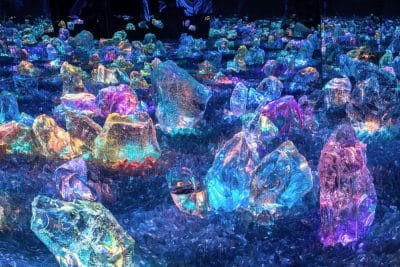 Große, farbenfrohe Kristalle und Felsen leuchten in kräftigem Blau, Rosa, Lila und Grün und schaffen eine magische Landschaft, die an das TeamLab Biovortex Kyoto erinnert - eine Verschmelzung von Technologie und Kunst in einer leuchtenden Sci-Fi-Fantasiewelt.