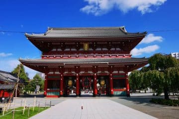 Großes traditionelles japanisches Tempeltor mit roten Säulen und verziertem Dach unter strahlend blauem Himmel. Bäume und Grünflächen umgeben das Bauwerk und machen es zu einem atemberaubenden Highlight auf jeder Reiseroute durch Japan. Ein gepflasterter Gehweg führt zum Eingang hinauf.