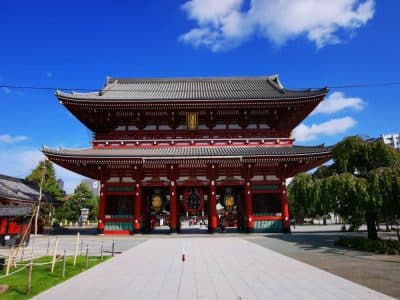 Großes traditionelles japanisches Tempeltor mit roten Säulen und verziertem Dach unter strahlend blauem Himmel. Bäume und Grünflächen umgeben das Bauwerk und machen es zu einem atemberaubenden Highlight auf jeder Reiseroute durch Japan. Ein gepflasterter Gehweg führt zum Eingang hinauf.