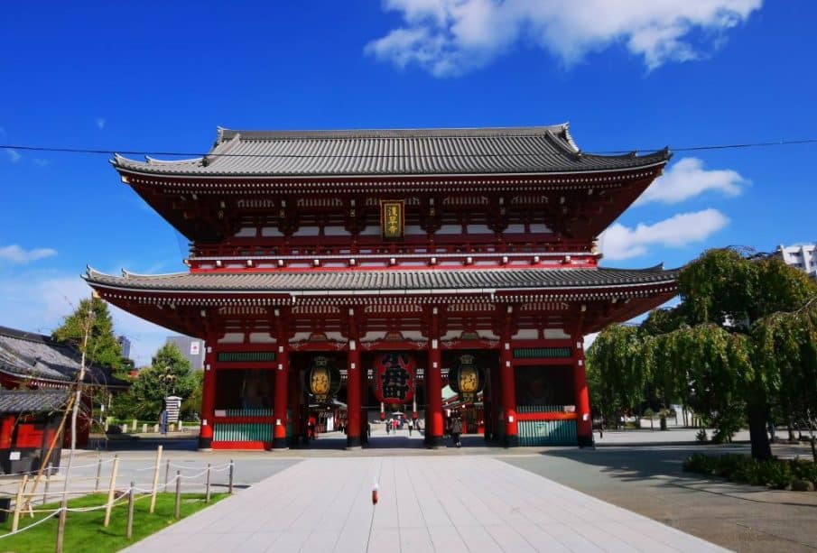 Großes traditionelles japanisches Tempeltor mit roten Säulen und verziertem Dach unter strahlend blauem Himmel. Bäume und Grünflächen umgeben das Bauwerk und machen es zu einem atemberaubenden Highlight auf jeder Reiseroute durch Japan. Ein gepflasterter Gehweg führt zum Eingang hinauf.