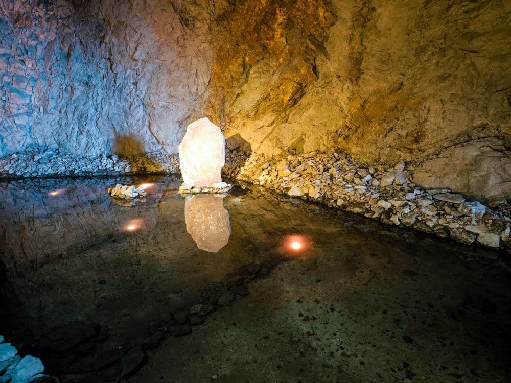 Ein glühender, kristallähnlicher Iwaya-Stein steht in einer unterirdischen Felsenhöhle und spiegelt sich im stillen Wasser, das den Boden teilweise überflutet. Kleine, schwache Lichter schwimmen auf der Wasseroberfläche und beleuchten die Höhlenwände.