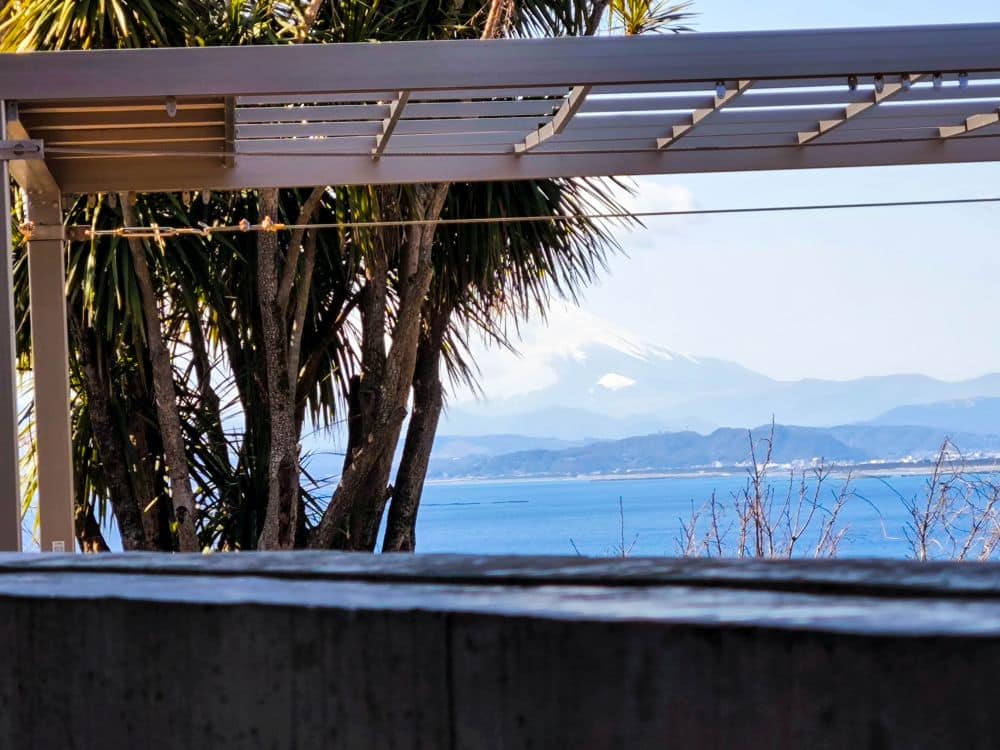 Ein malerischer Blick auf den Berg Fuji in der Ferne, teilweise von Wolken verdeckt, durch Palmen und eine Pergola hindurch, mit dem blauen Meer und den Hügeln im Vordergrund - und der berühmten Enoshima-Meereskerze am Horizont.